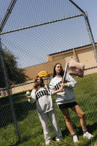 Two women posing in a baseball field wearing matching light gray sweatpants and sweatshirts with 'BASEBALL' printed in black. One holds a baseball bat and the other holds a baseball, with a chain-link fence and grassy field in the background.