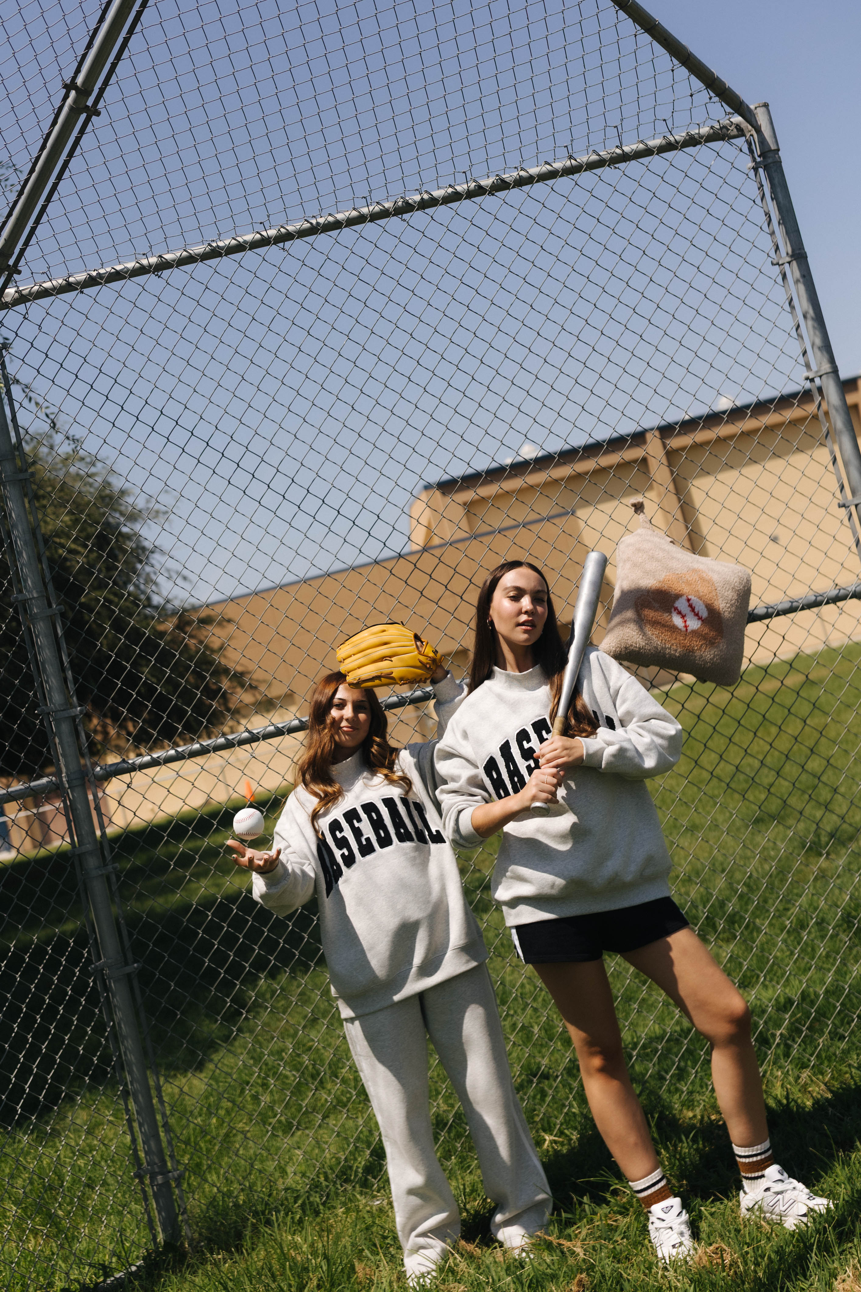 Two women posing in a baseball field wearing matching light gray sweatpants and sweatshirts with 'BASEBALL' printed in black. One holds a baseball bat and the other holds a baseball, with a chain-link fence and grassy field in the background.