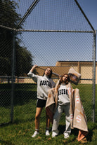 Two women posing outdoors in front of a chain-link baseball fence, wearing matching light gray sweatshirts with 'BASEBALL' printed across the chest. One holds a baseball bat, and both have beige blankets with baseball-themed prints draped over their arms.