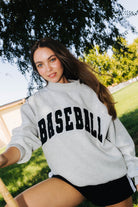 A woman outdoors wearing a light gray sweatshirt with 'BASEBALL' printed across the front in black arched lettering, paired with black shorts.
