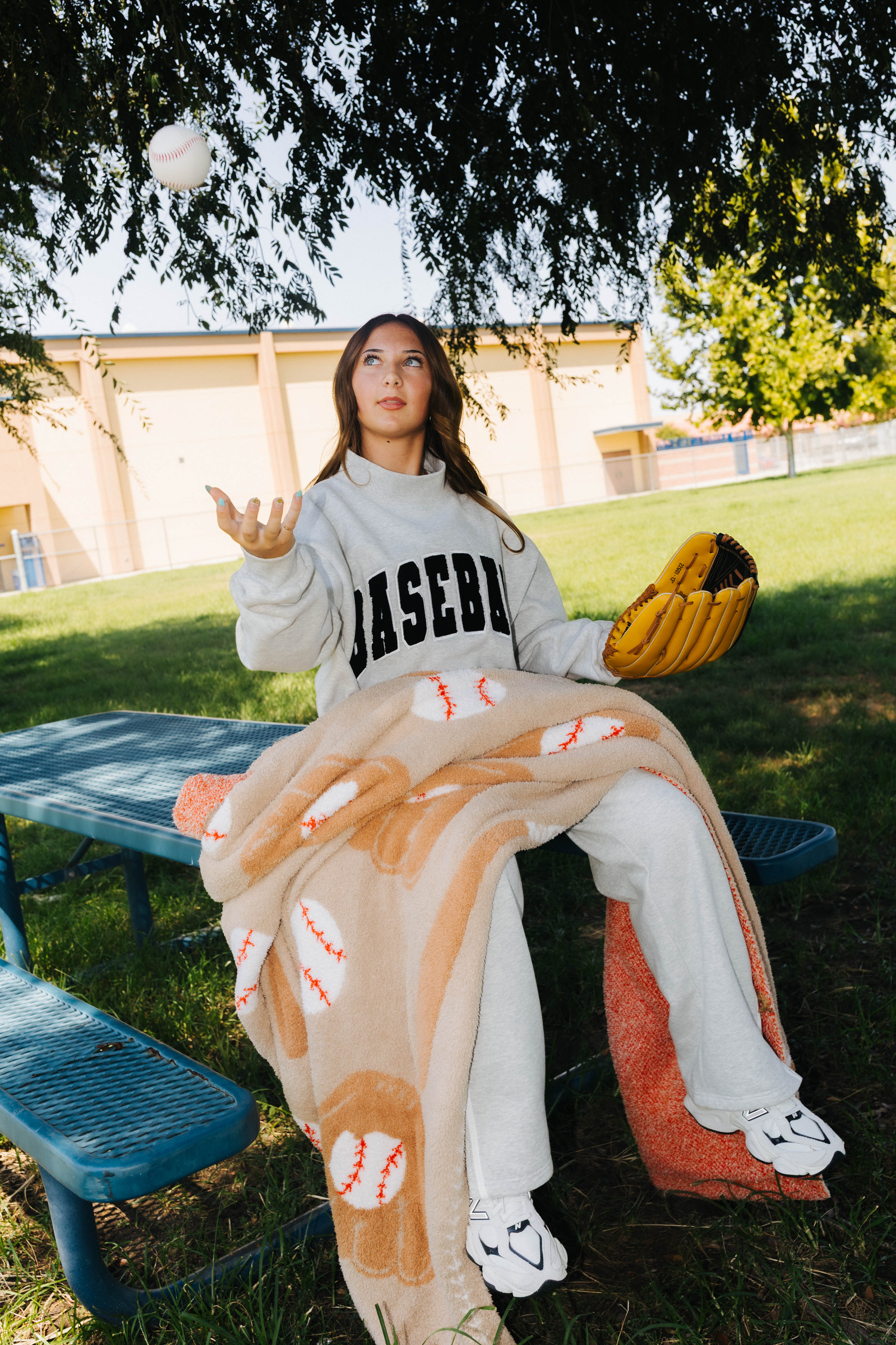 A woman sitting on a blue picnic table outdoors under a tree, wearing a light gray sweatshirt with 'BASEBALL' printed in black, gray sweatpants, a tan and brown baseball-themed blanket, and holding a yellow baseball glove.