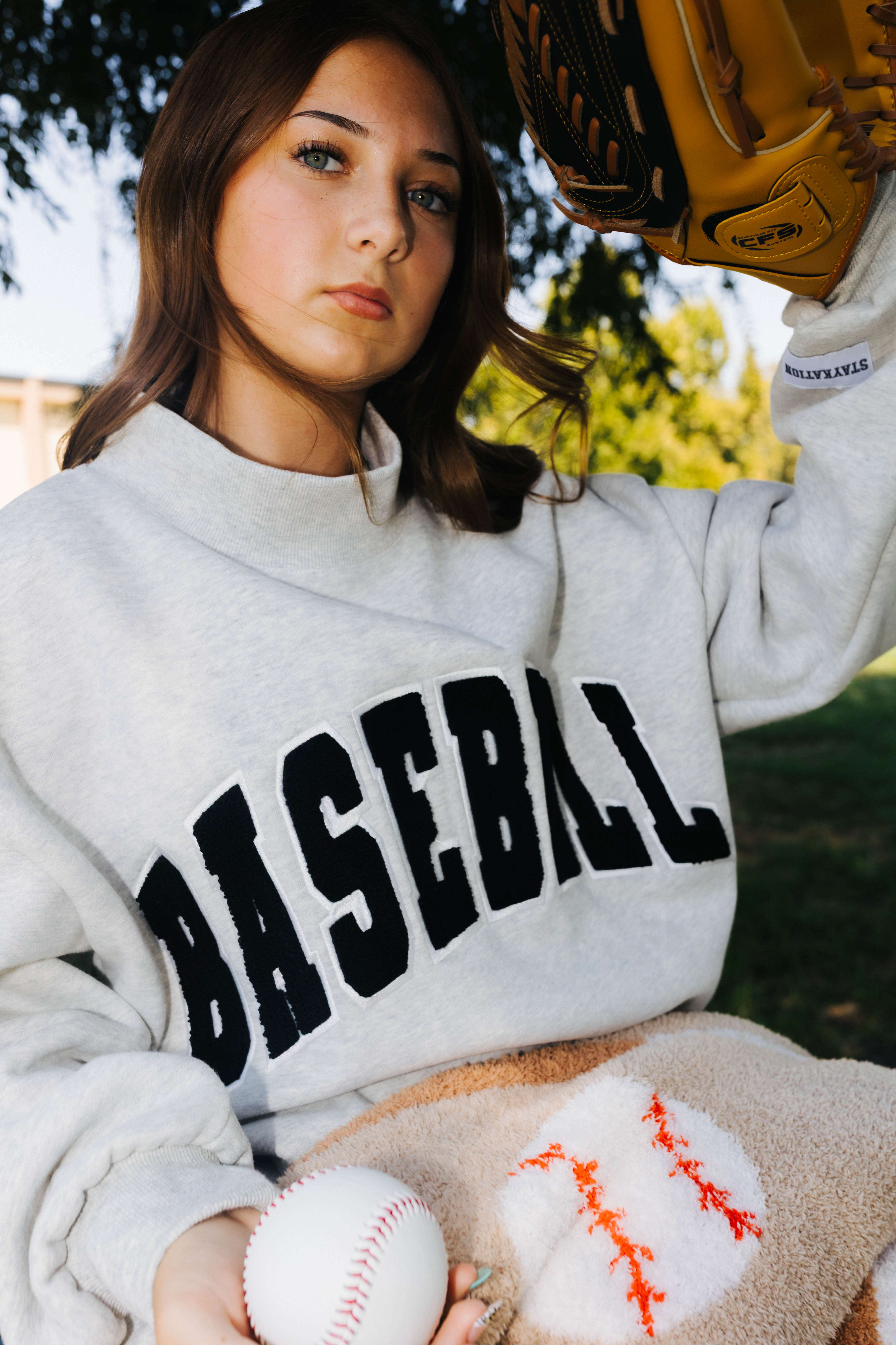 A woman outdoors wearing a light gray sweatshirt with 'BASEBALL' printed across the chest in black varsity letters. She is holding a baseball and a tan baseball glove, with trees and grass in the background.