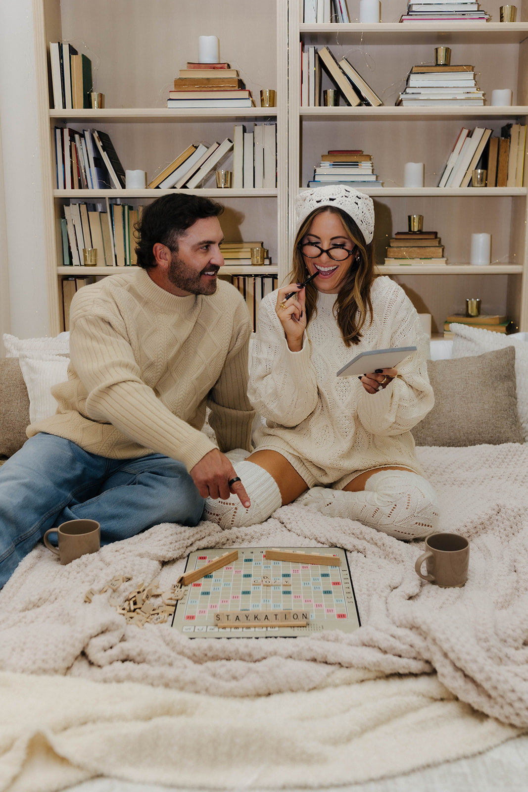 Man and woman sitting on a bed with a blanket, surrounded by books and a tablet.