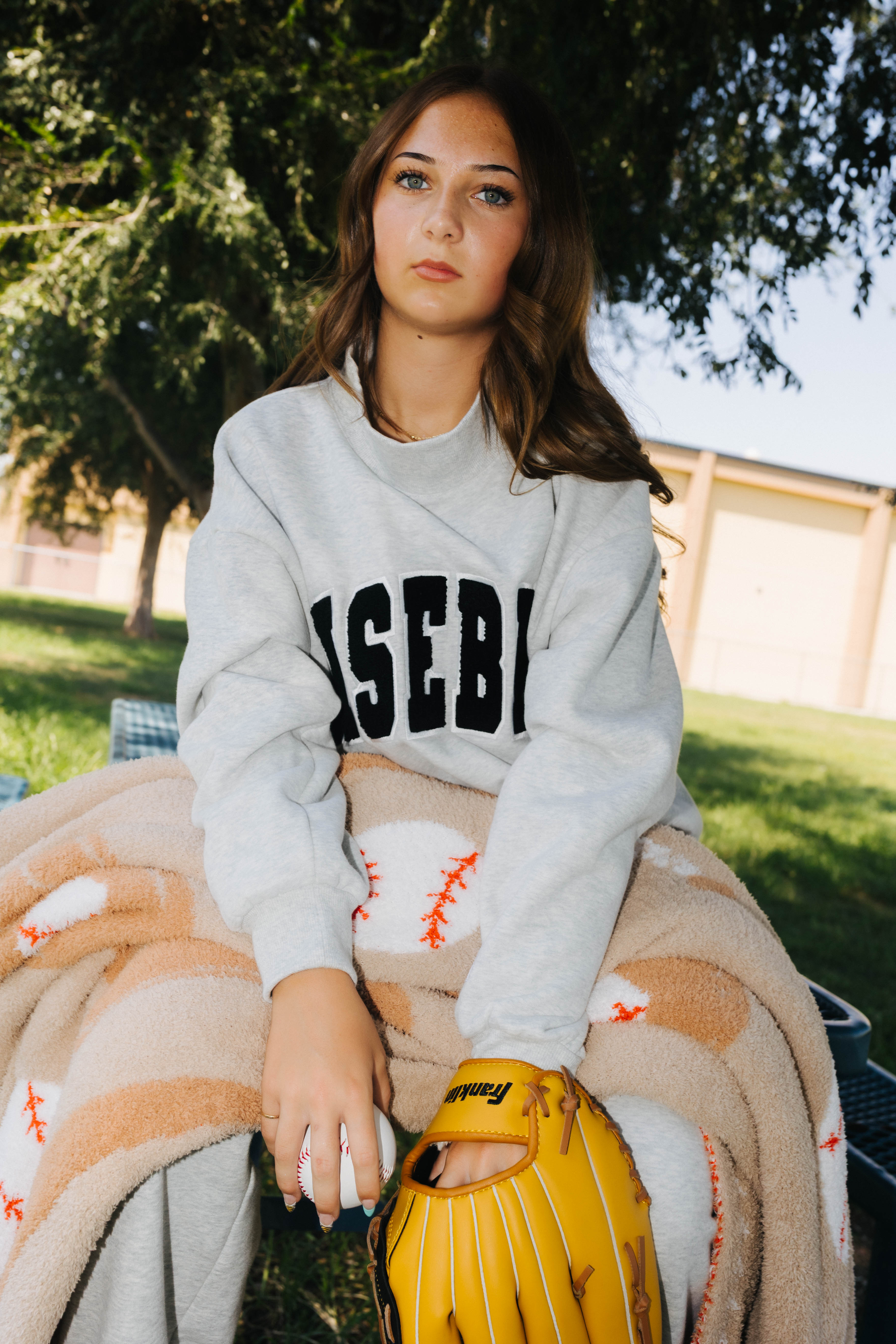 A woman sitting outdoors on a blanket, wearing a light gray sweatshirt with 'BASEBALL' printed in black across the chest, holding a yellow baseball glove.