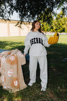 A woman stands outdoors on grass under a tree wearing a light gray baseball-themed sweatshirt and sweatpants set, holding a tan baseball-patterned blanket and a yellow glove.
