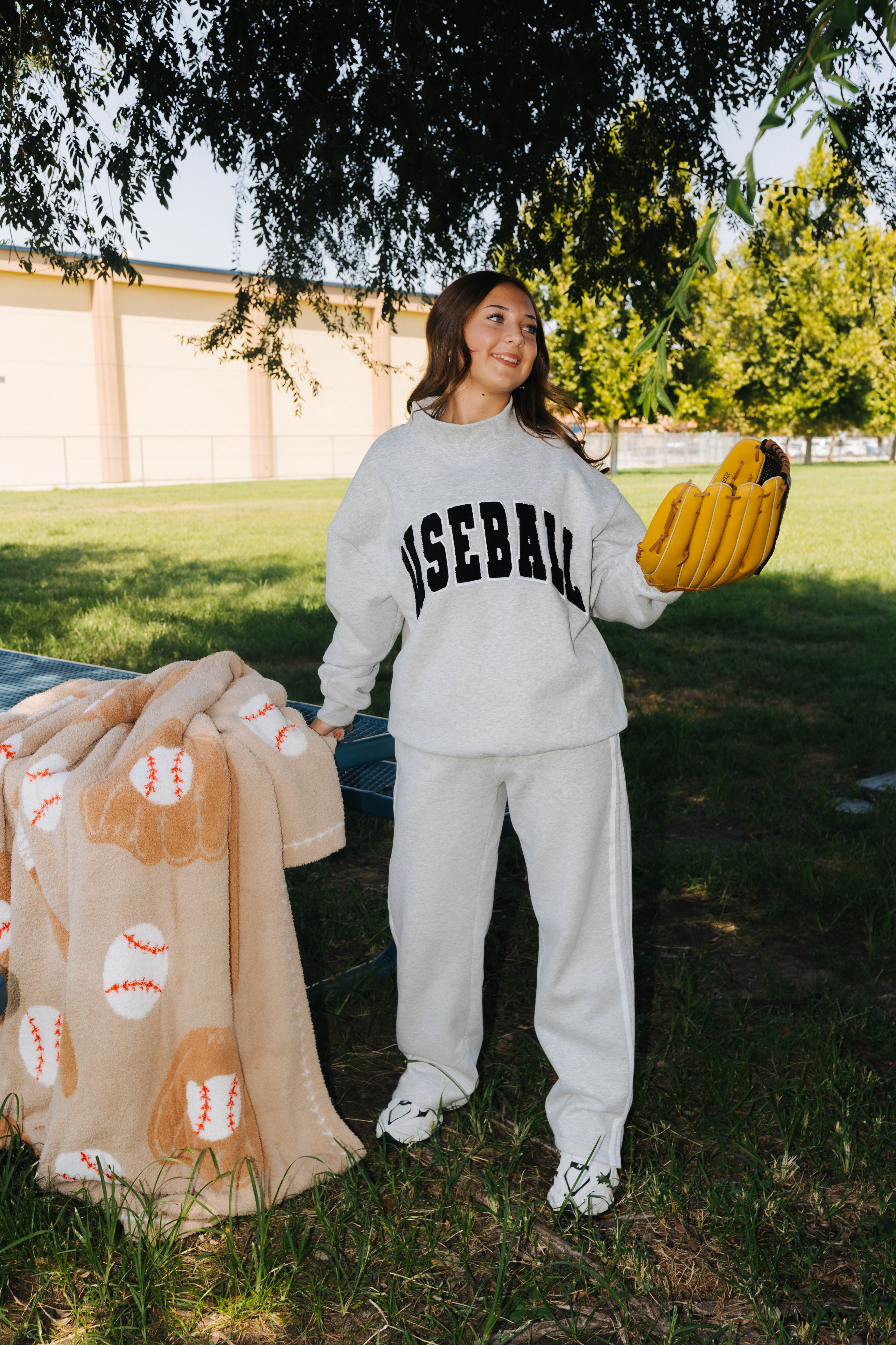 A woman stands outdoors on grass under a tree wearing a light gray baseball-themed sweatshirt and sweatpants set, holding a tan baseball-patterned blanket and a yellow glove.