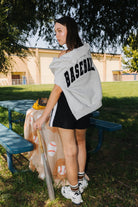 girl holding a baseball bat wearing a baseball sweatshirt draped around her neck with a baseball blanket lying on the table in front of her