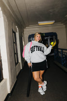 A woman stands in a hallway wearing a light gray hoodie with 'SOFTBALL' printed in black across the chest, paired with a black shorts, white sneakers, and a pink quillow. She is holding a baseball bat and softball.