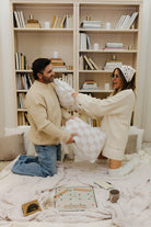 Man and woman in a cozy room with bookshelves and a blanket, holding pillows.