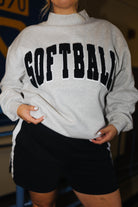A person wearing a light heather gray mock-neck sweatshirt with 'SOFTBALL' printed across the chest in large black varsity-style letters, paired with black athletic shorts.