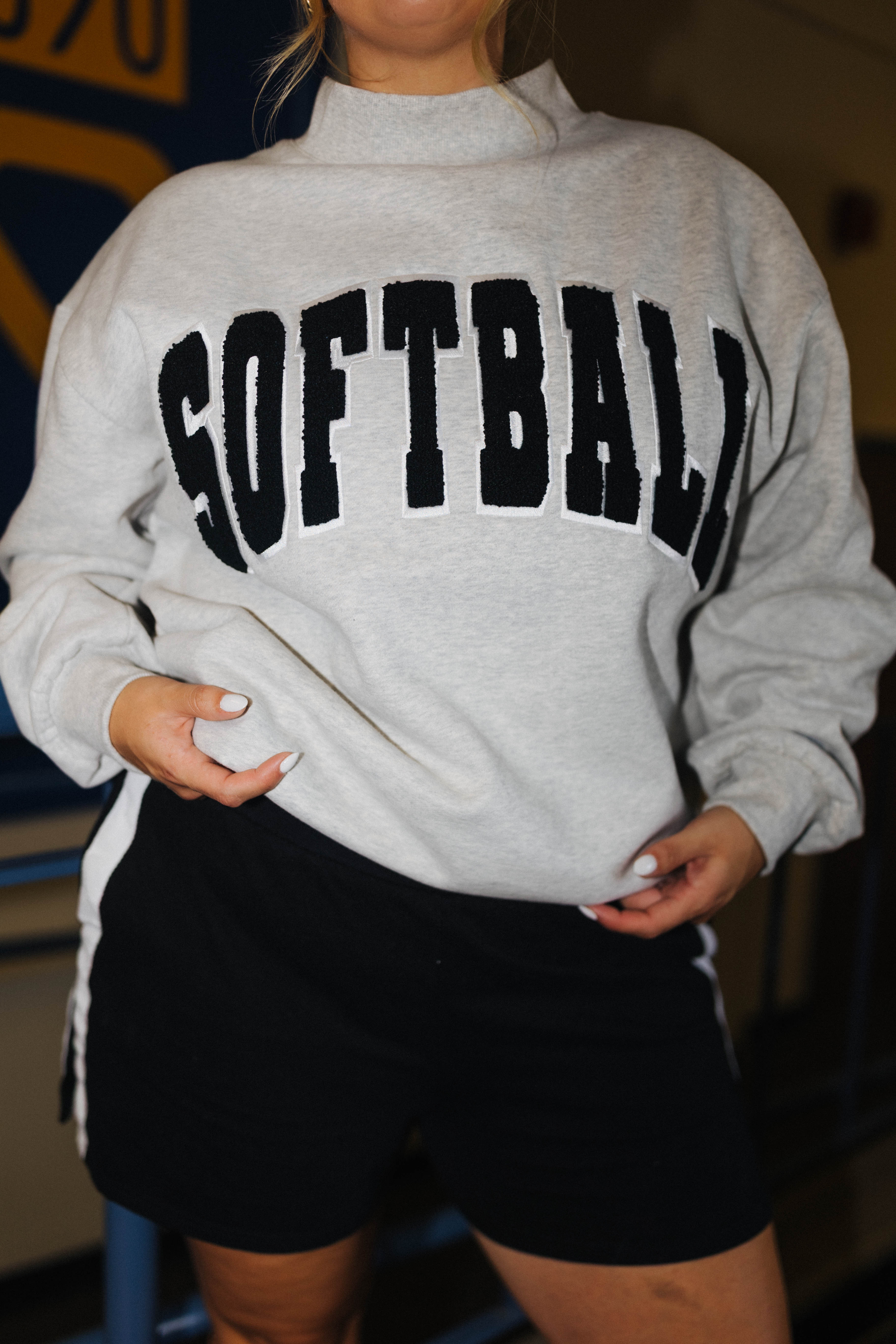 A person wearing a light heather gray mock-neck sweatshirt with 'SOFTBALL' printed across the chest in large black varsity-style letters, paired with black athletic shorts.