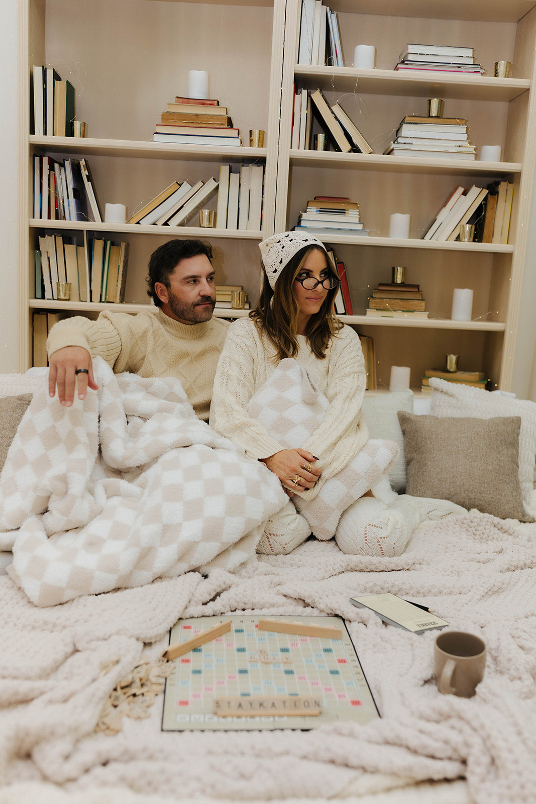 Two people sitting on a couch under a blanket with a bookshelf in the background