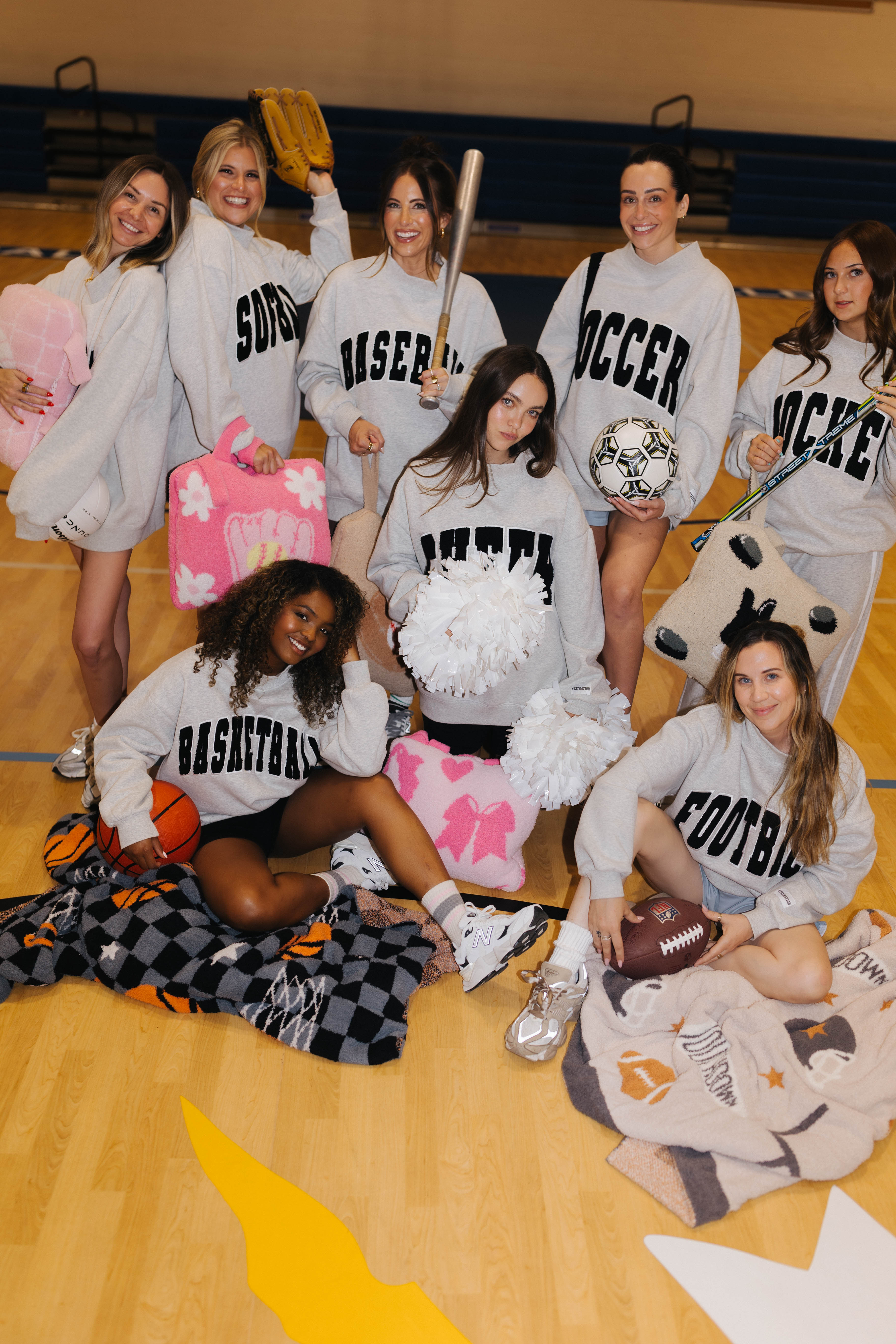 A group of women wearing matching gray athletic sweatshirts with team lettering, posed together on a wooden gymnasium floor with sports balls, plush quillows and blankets.
