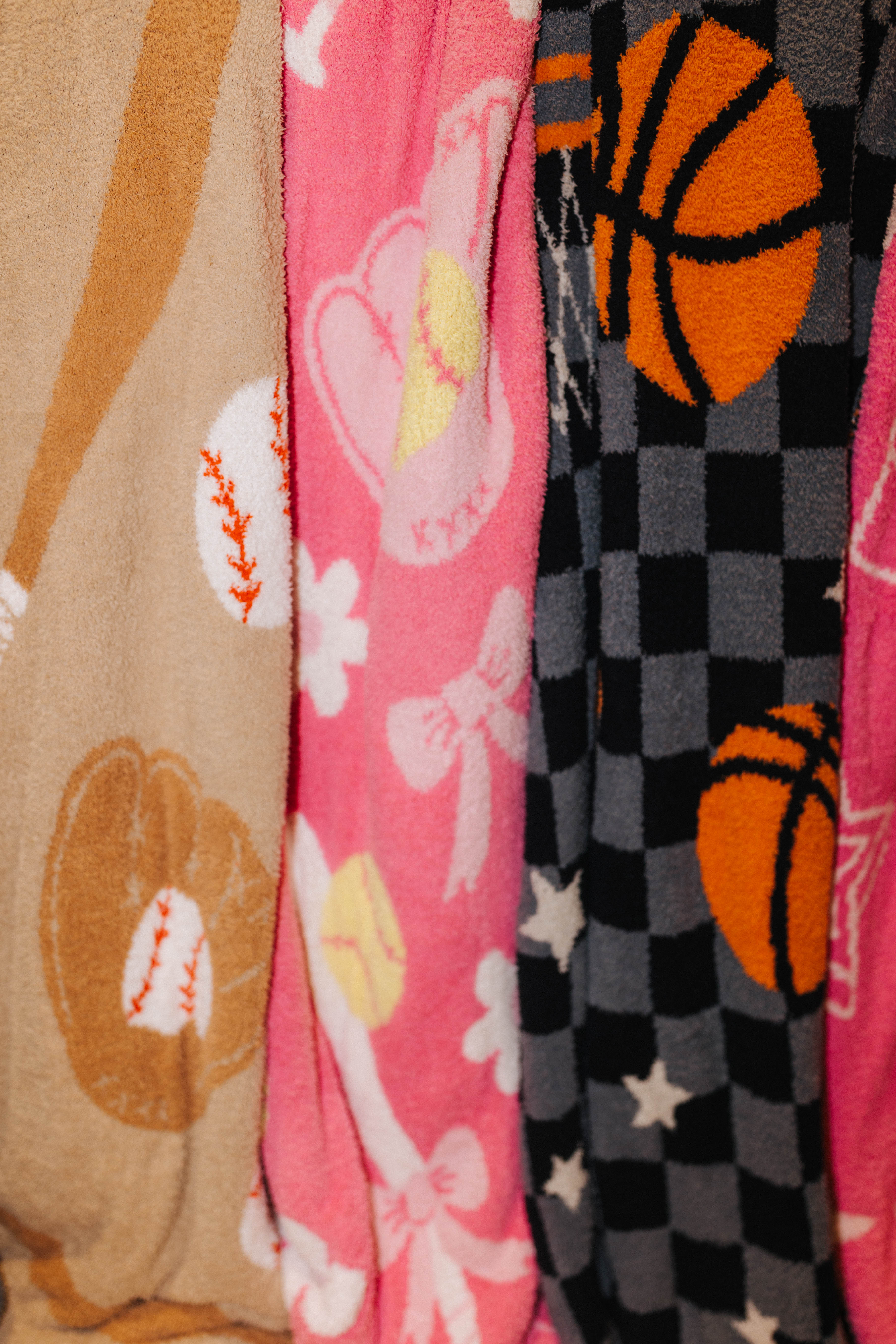 Close-up of three patterned fabric items hanging side by side, including a tan blanket with baseball motifs, a pin blanket with bows and softballs, and a dark gray/black checkered fabric with orange basketballs and white stars.