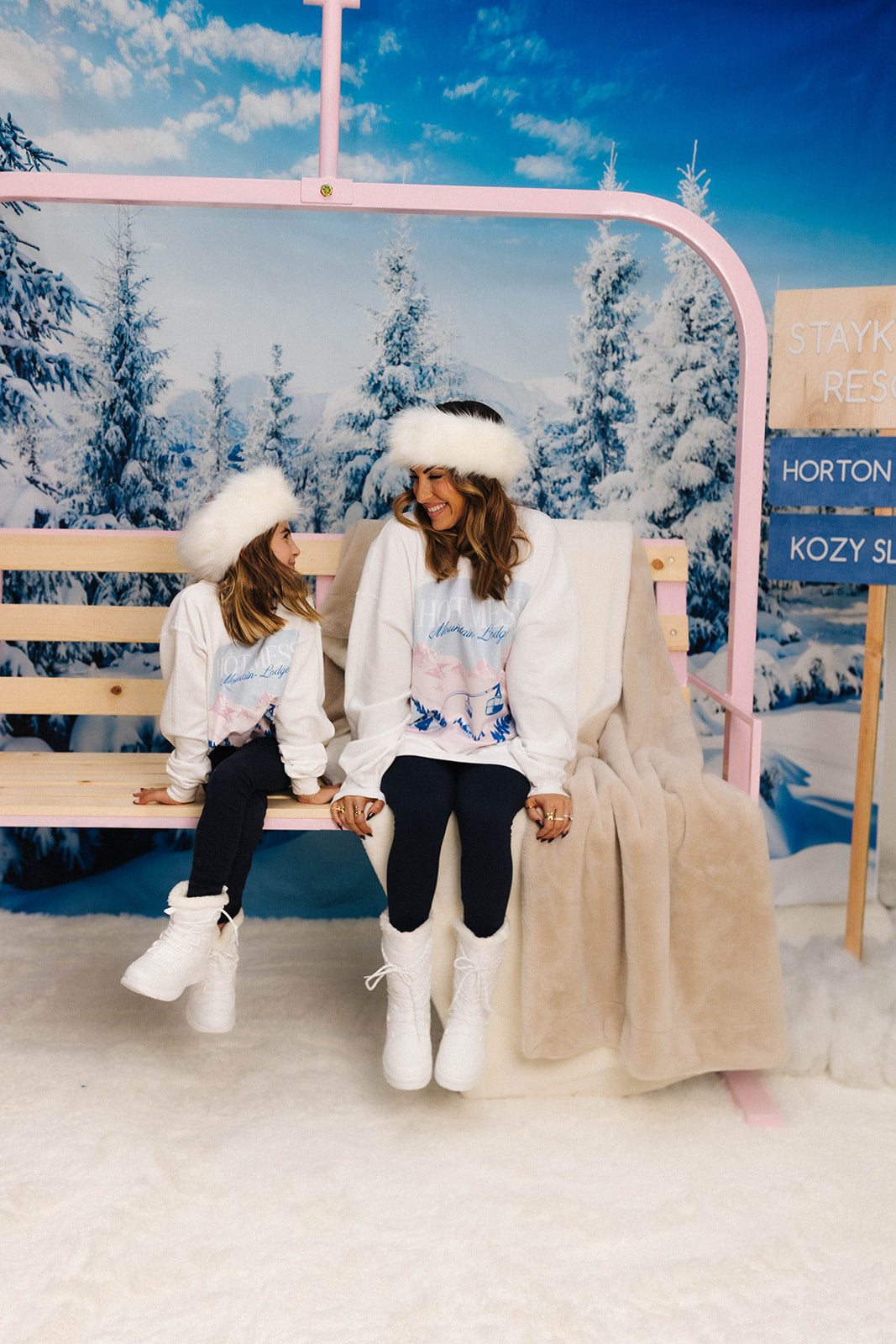 mother and daughter smiling at each other wearing matching ski sweatshirts on a ski slope backdrop 