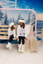 mother and daughter smiling at each other wearing matching ski sweatshirts on a ski slope backdrop 