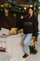 Woman in a brown sweatshirt with text standing in a room decorated with Christmas trees and gold stars.