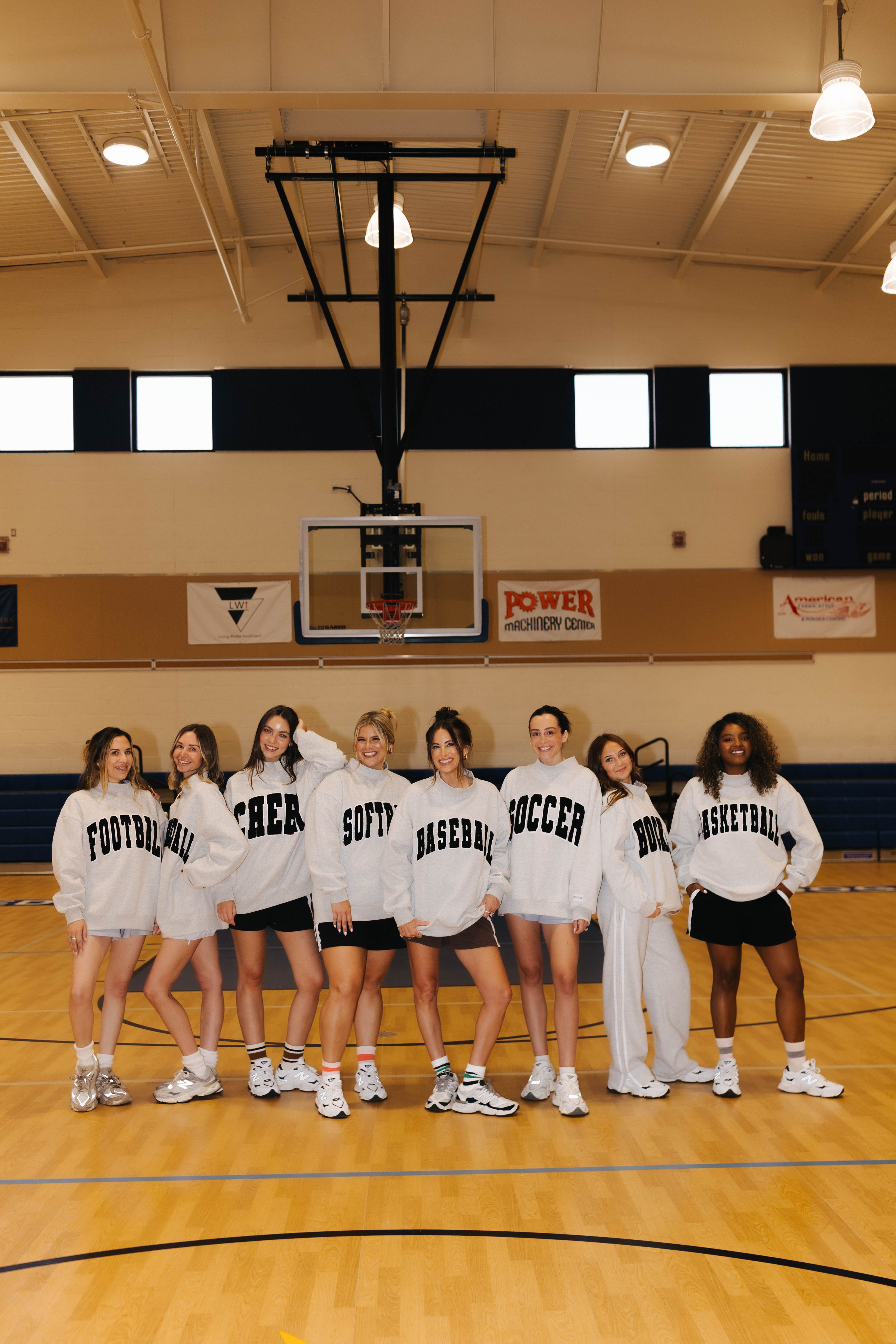 A group of nine women posing together on a wooden indoor basketball court, wearing matching white crewneck sweatshirts with different sports words printed on the front (such as Football, Cheer, Softball, Baseball, Soccer, and Basketball).