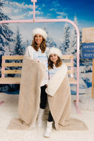 Two people wrapped in a blanket in a snowy scene with a pink structure and trees.