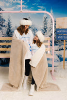 Two people wrapped in a blanket in a snowy landscape with a pink structure and signs in the background.