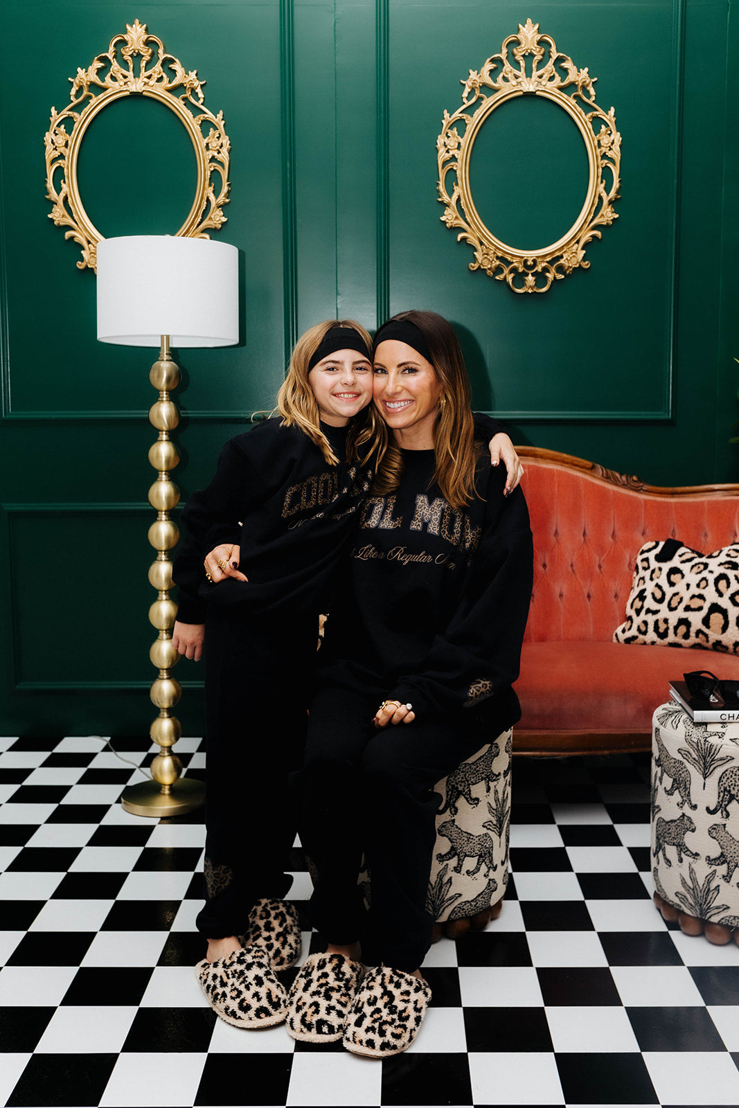 Two women posing together in a stylish room with gold mirrors, a red couch, and checkered floor.