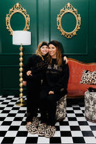 Two women posing together in a stylish room with gold mirrors, a red couch, and checkered floor.