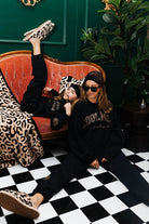 Two people sitting on a checkered floor with a red couch and green wall in the background.