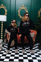 Two people in matching black outfits with leopard print details standing in a room with green walls and decorative mirrors.