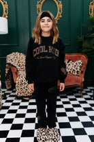 Person wearing a black sweatshirt with 'Cool Kid' text in a room with checkered floor and decorative wall.