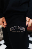 Black sweatpant with 'Cool Mom Not a Regular Mom' text held by a person.