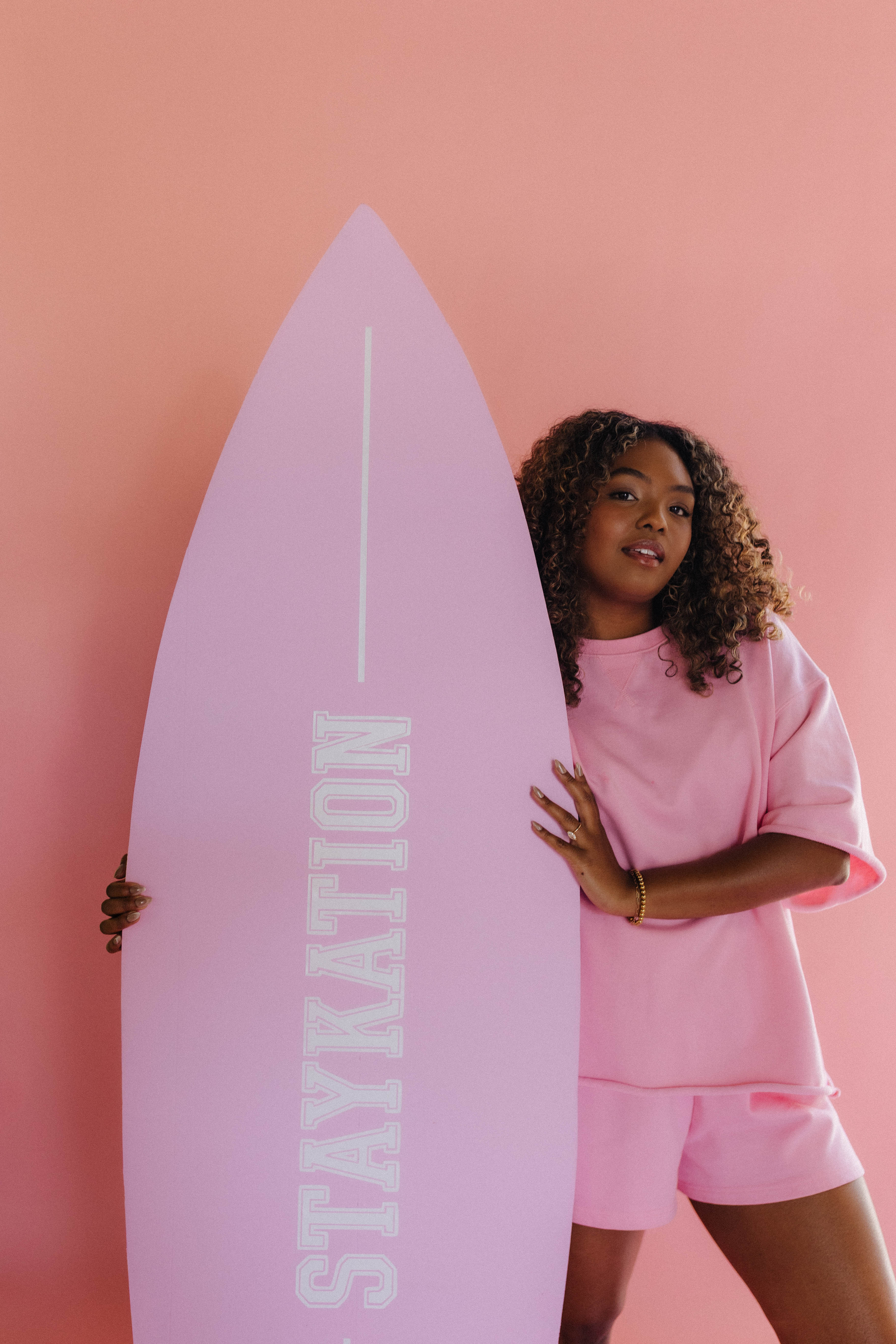 Person holding a pink surfboard with 'STAYKATION' text against a pink background