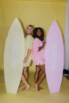 Two women holding surfboards with a yellow background
