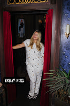 A woman wearing a white short-sleeve pajama set with a black and gold bat pattern stands in a doorway framed by red curtains. The text overlay at the bottom left reads 'ENSLEY IN 2XL'.