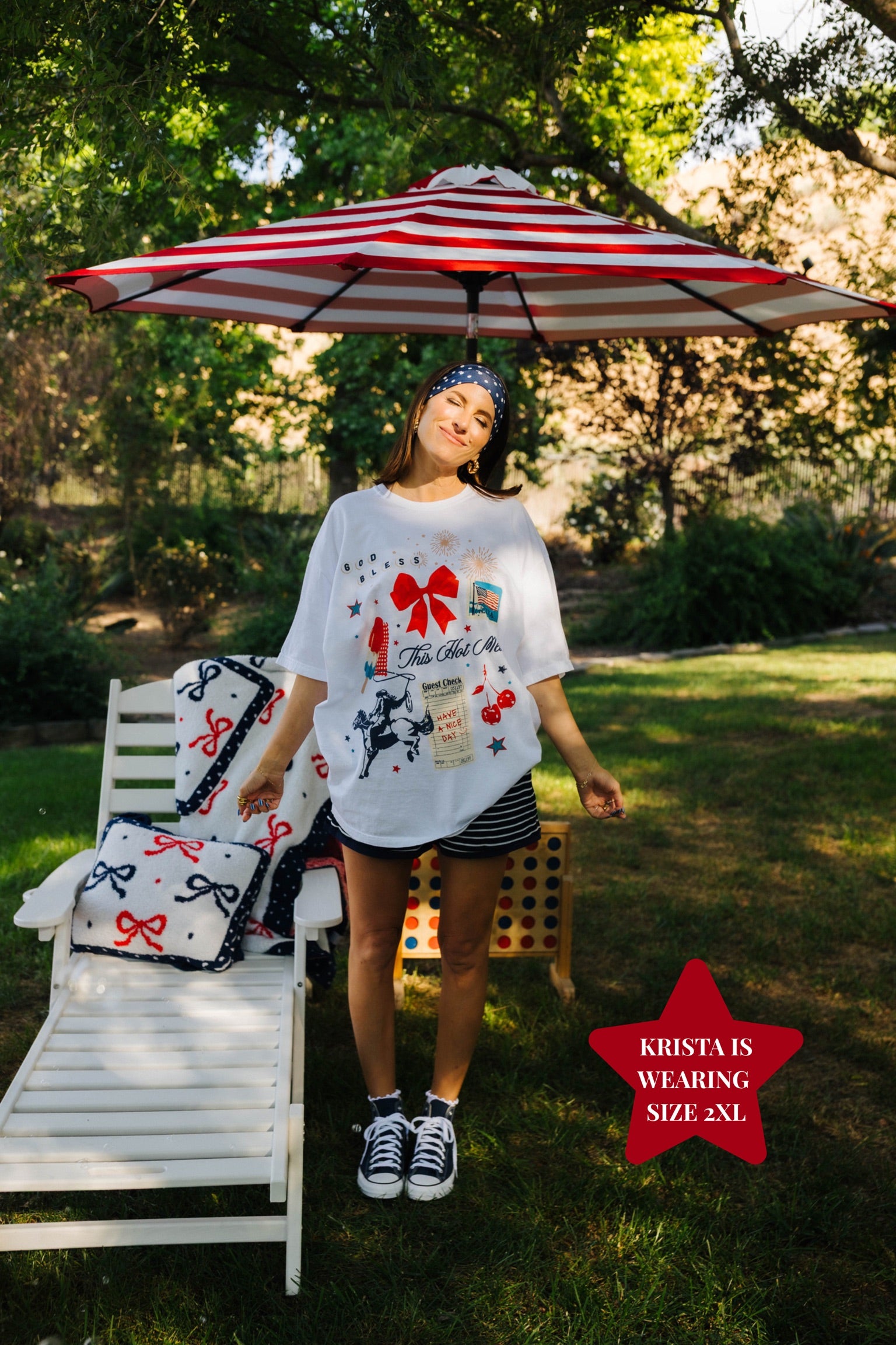 Woman standing under a red and white striped umbrella with a garden chair and decorative pillows in the background.