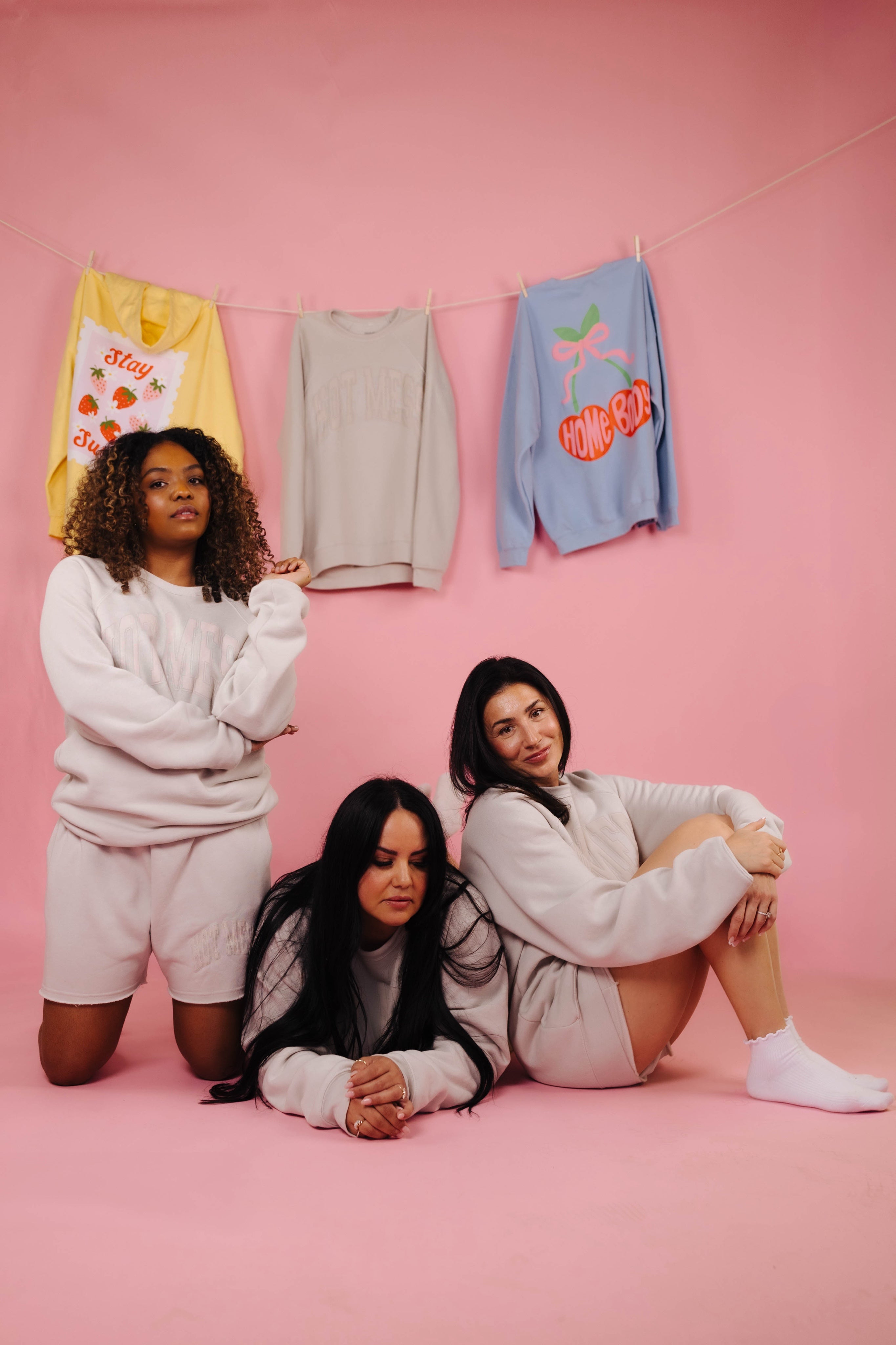 Three women posing on a pink background with colorful sweatshirts hanging on a line.