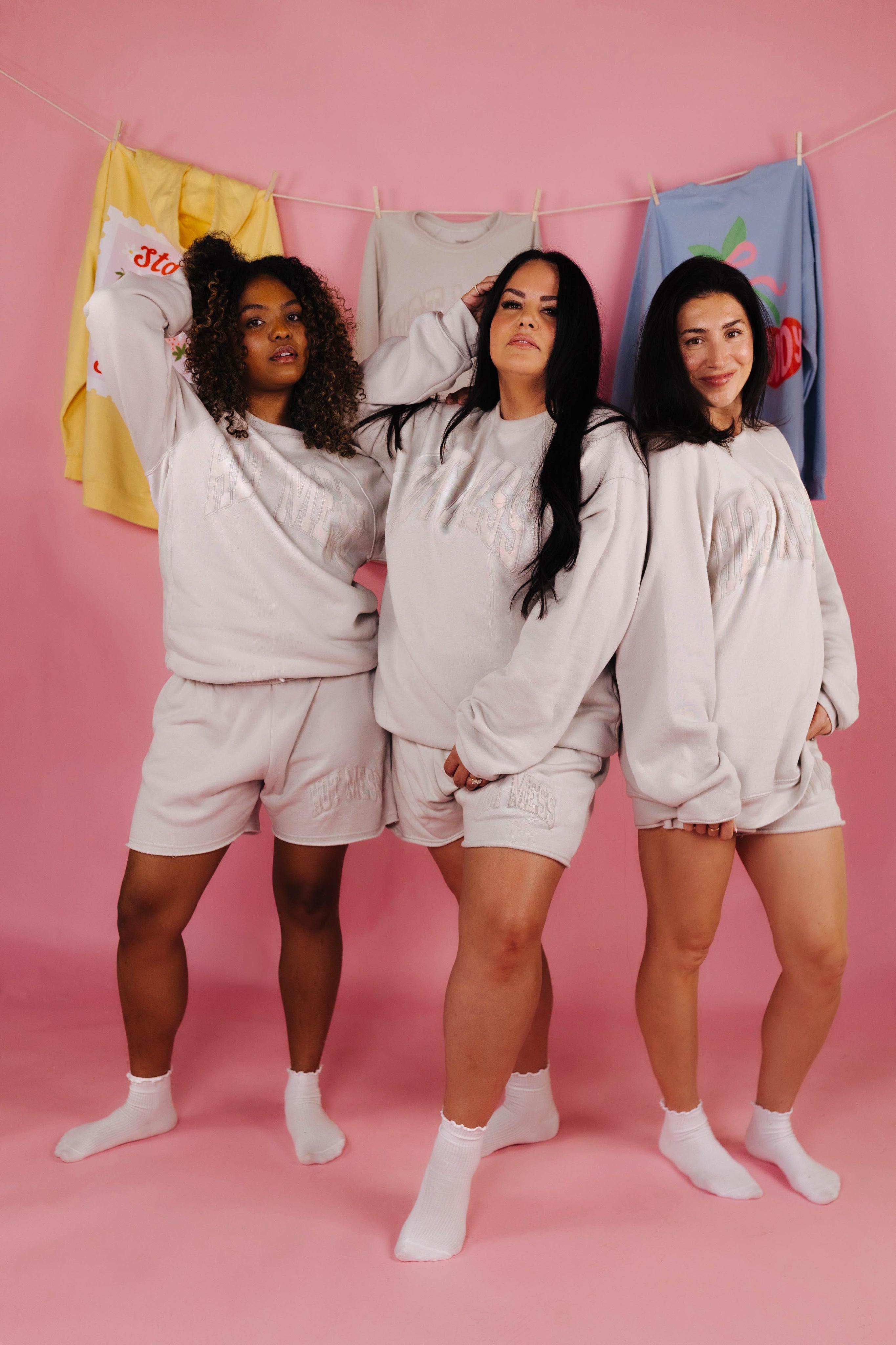 Three women wearing matching tan hot mess loungewear sets against a pink background with colorful shirts hanging on a line.