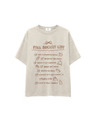 A light beige oversized t-shirt with a brown graphic print titled 'FALL BUCKET LIST' featuring various autumn-themed checklist items, shown against a plain white background.