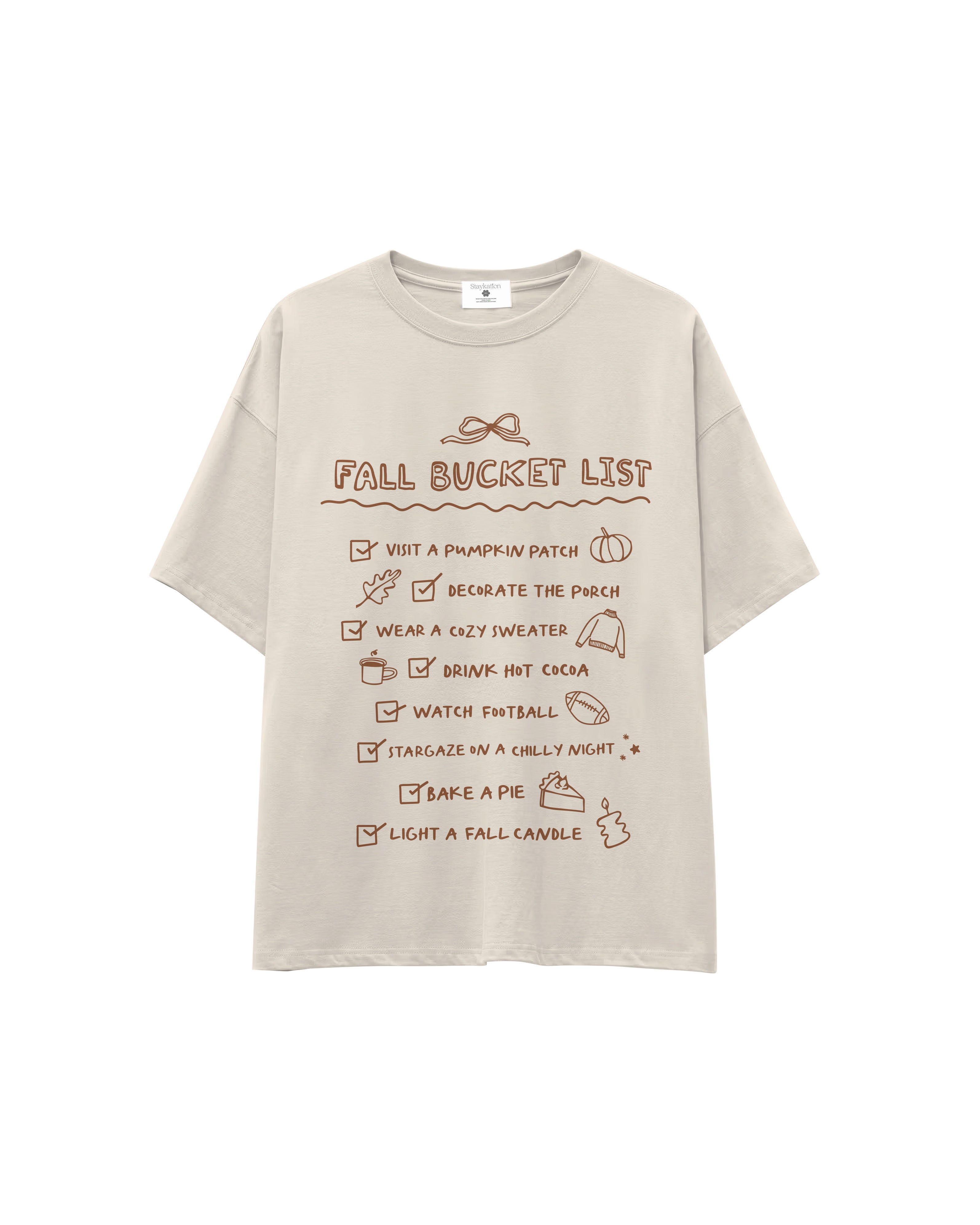 A light beige oversized t-shirt with a brown graphic print titled 'FALL BUCKET LIST' featuring various autumn-themed checklist items, shown against a plain white background.