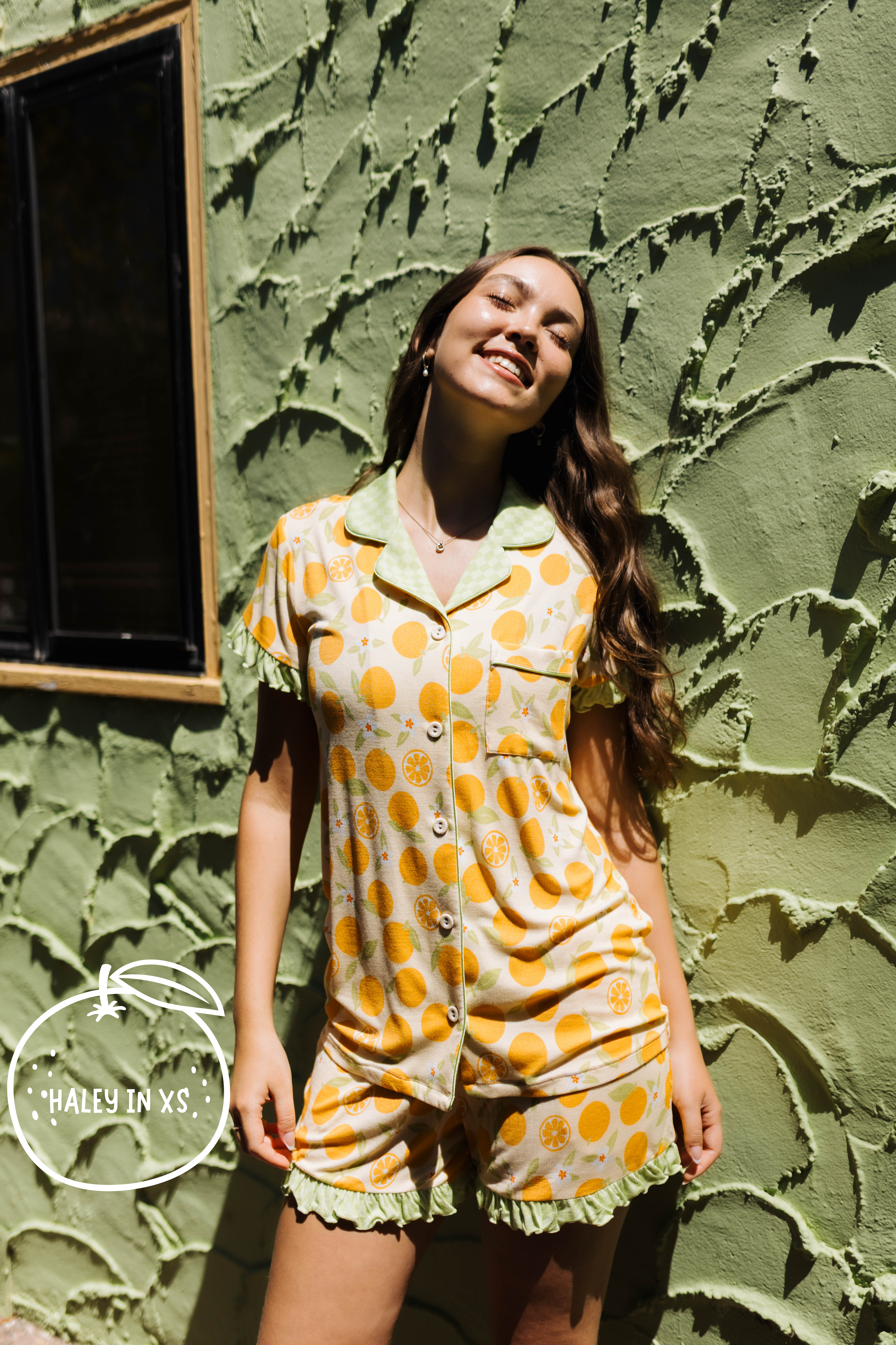 A woman wearing a short-sleeve button-front pj matching ruffled shorts, featuring orange citrus fruit print. She is posing outdoors against a textured olive green stucco wall.