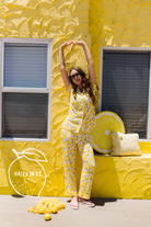 A woman modeling a yellow lemon-print pajama set stands in front of a textured yellow wall decorated with lemon props. She is wearing sunglasses and flip-flops, with a brand watermark 'HALEY IN XS' visible in the lower-left corner.