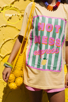 A person wearing a beige graphic T-shirt with a colorful 'HOTMess express' print, styled with a yellow mesh tote bag, a chunky multicolor beaded necklace, and pink shorts against a textured yellow wall.