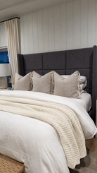 A styled bedroom scene showing a bed with a tall dark gray upholstered headboard, white bedding, beige pillows, and a cream textured throw blanket. The wall behind the bed has vertical white paneling.