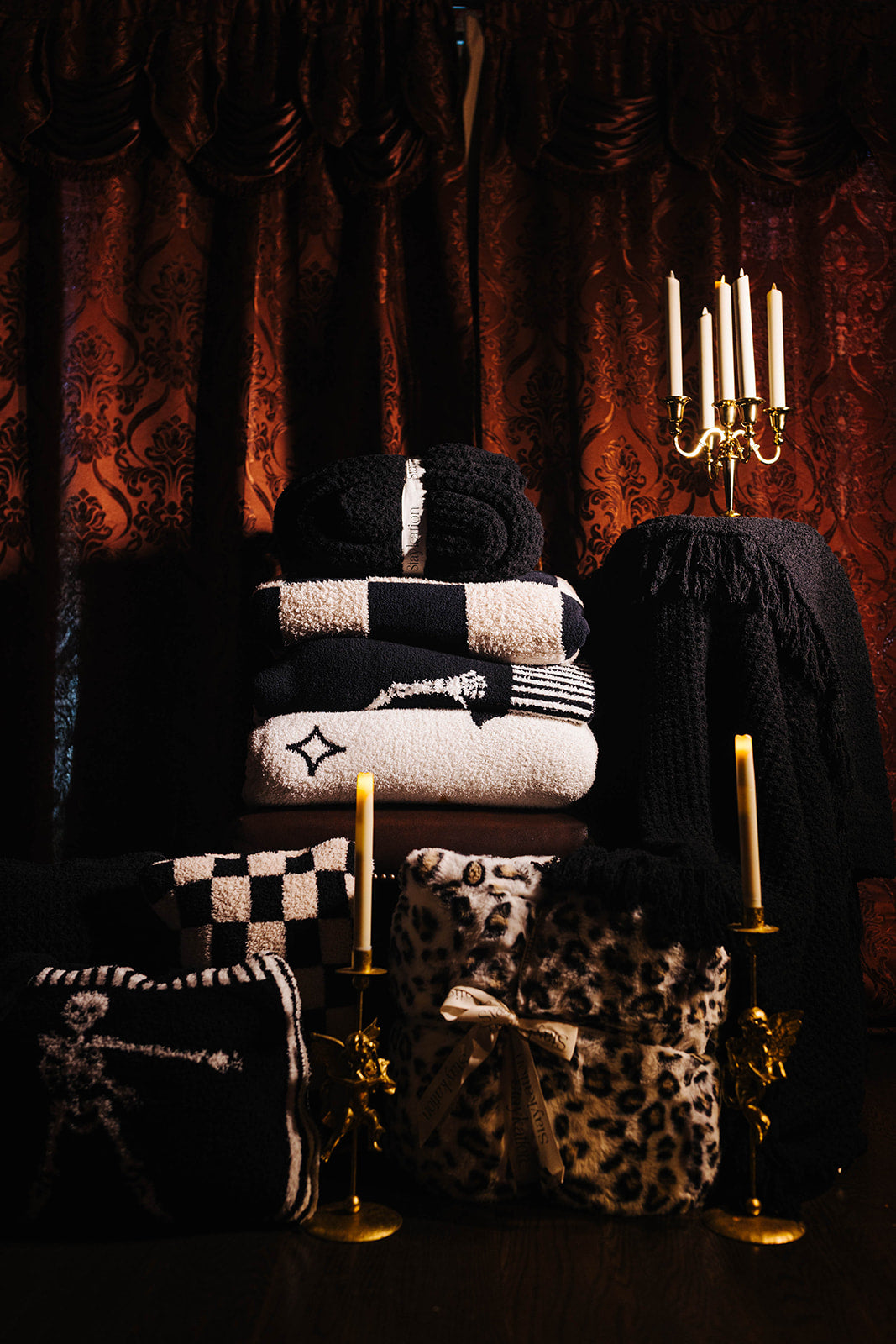 A styled collection of black and cream knit blankets, throw pillows, and a leopard-print fabric item arranged on a dark wooden surface against a backdrop of ornate dark red curtains, with gold candlesticks as props.
