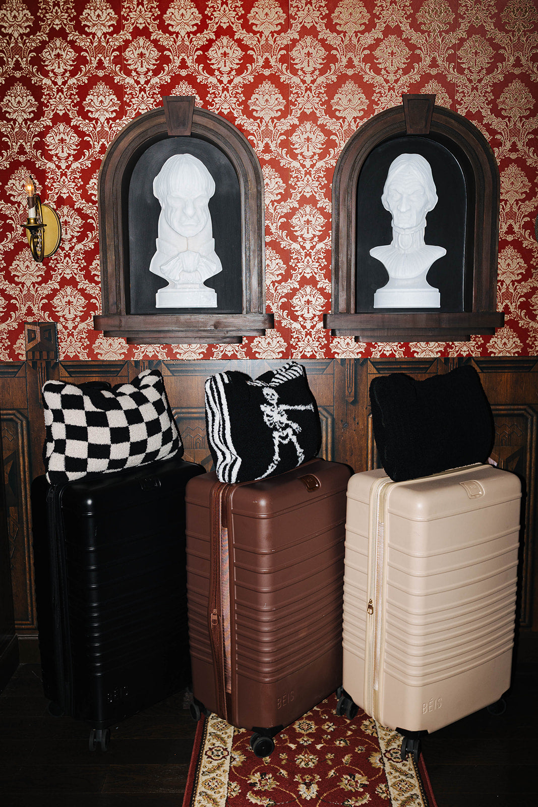 Three hard-shell suitcases in black, brown, and cream are arranged on a patterned red rug in front of ornate red damask wallpaper and two dark wood arched frames containing white bust sculptures.