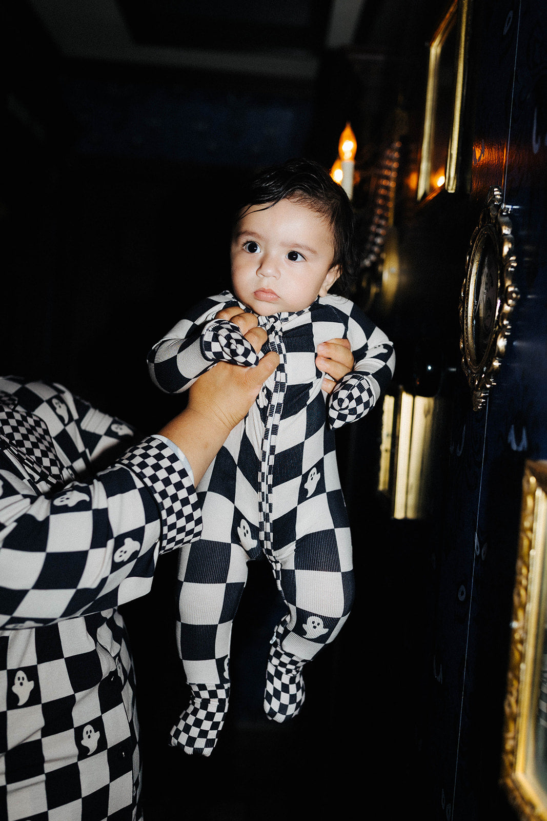 A baby wearing a black and sand checkered footed onesie is held up by an adult wearing a matching checkered shirt in a dark interior setting.