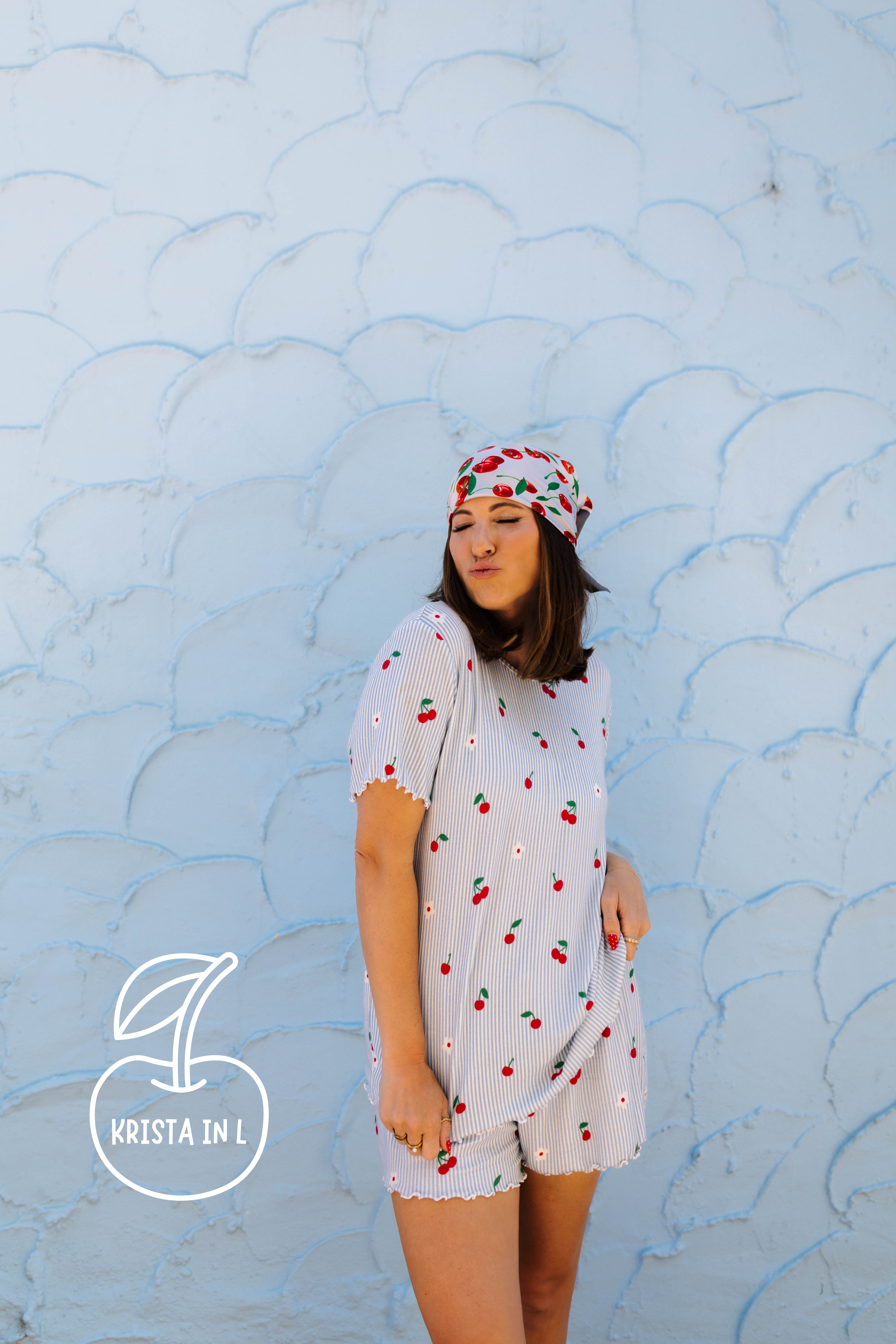 A woman wearing a white cherry-patterned pj and matching headscarf stands against a light blue textured wall. The brand logo 'Krista in L' with an cherry icon is visible in the bottom-left corner.