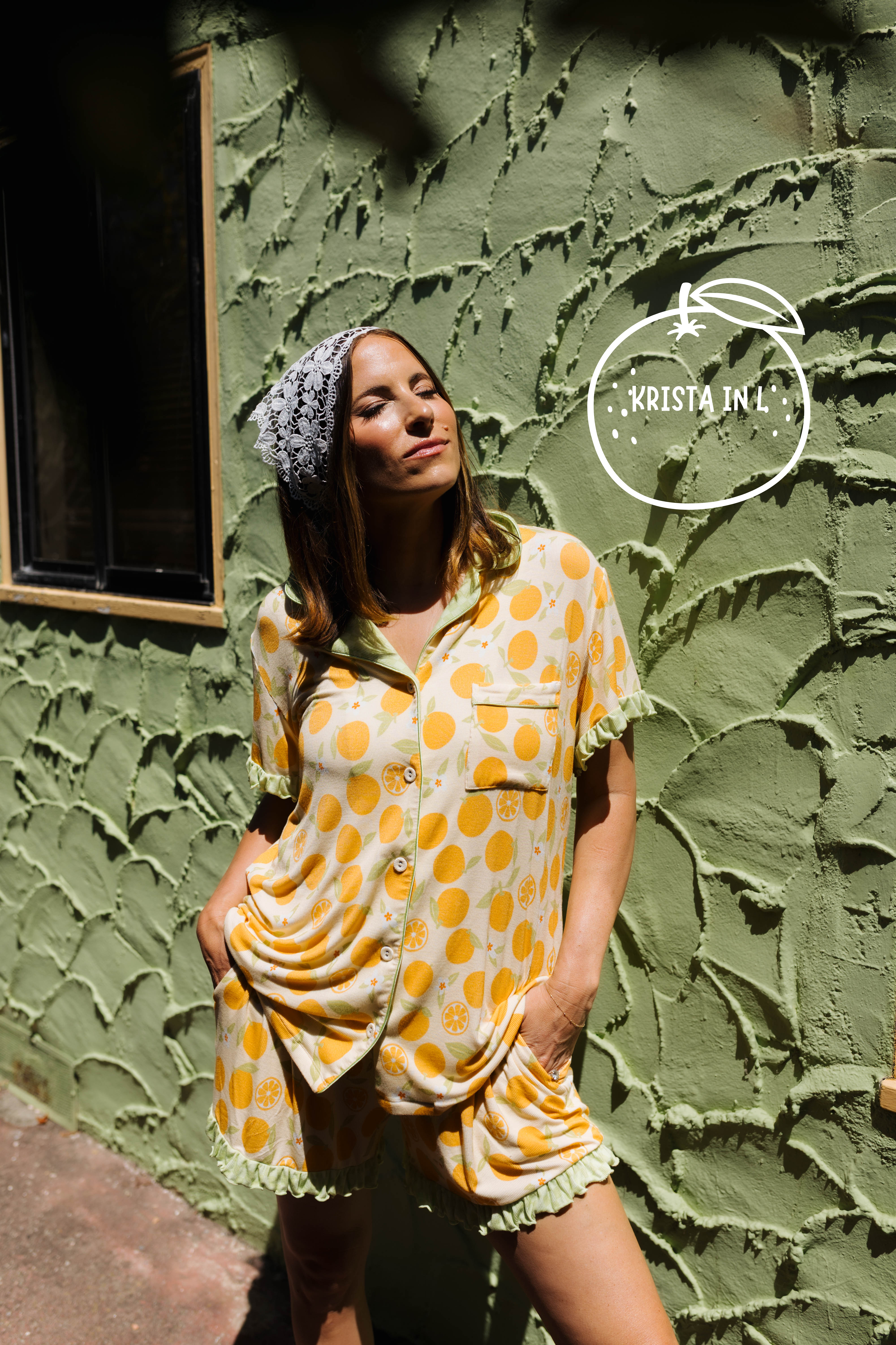 A woman wearing an orange pajama set with a matching stands against a textured olive green stucco wall. A white 'KRISTA IN A' logo watermark is visible in the upper right.