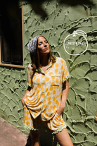 A woman wearing an orange pajama set with a matching stands against a textured olive green stucco wall. A white 'KRISTA IN A' logo watermark is visible in the upper right.