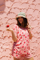 A woman wearing a pink strawberry-print tunic top and matching pj shorts stands against a textured pink wall, holding a strawberry. A 'Krista in L' logo watermark is visible in the lower-left corner.