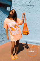 A woman wearing an oversized peach-orange graphic t-shirt dress with 'Home Body' text and orange illustrations, holding a mesh bag of oranges against a textured light blue wall.